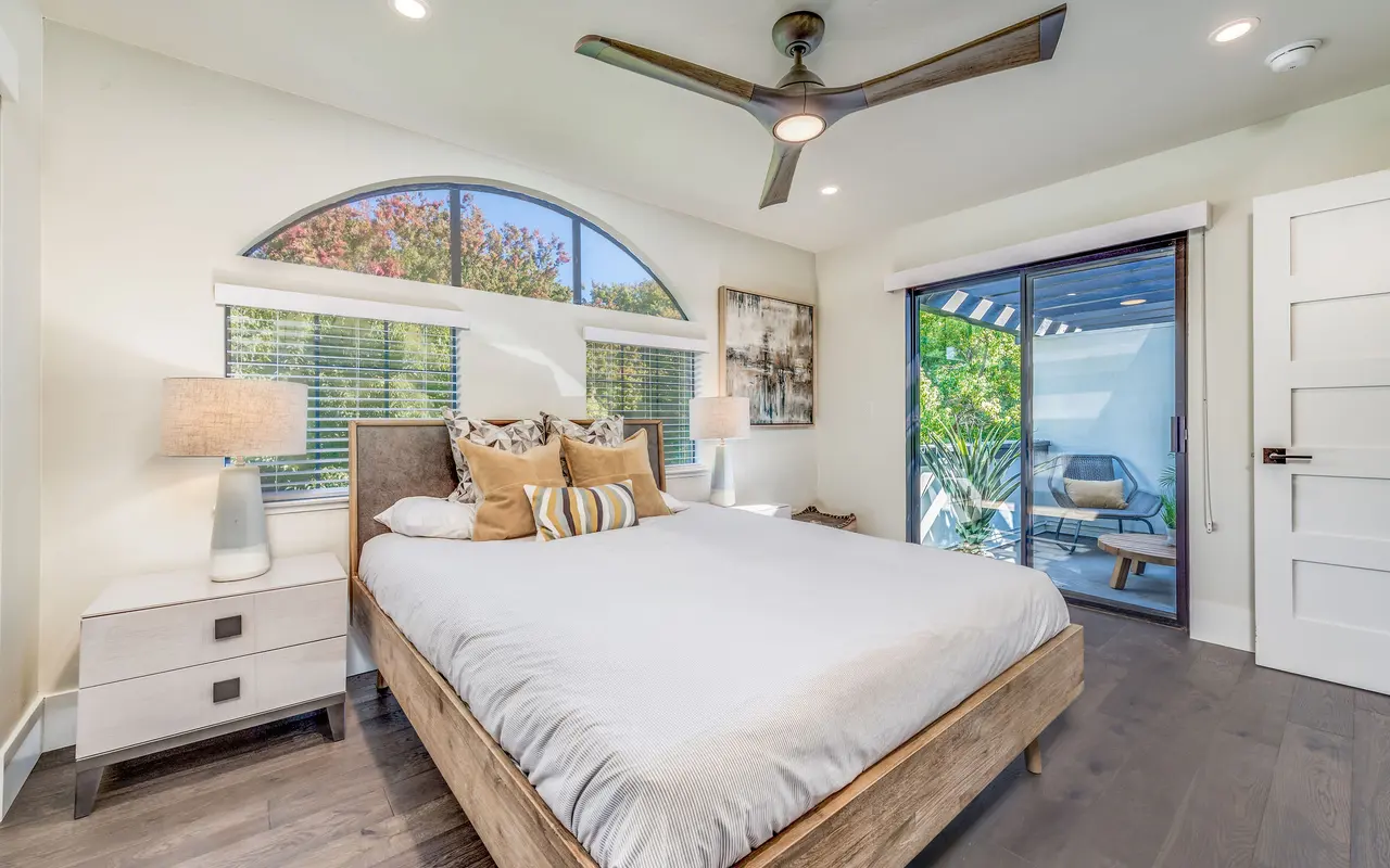 A modern bedroom featuring a queen-sized bed with white bedding, decorative pillows, two stylish bedside lamps, and a large window with an arched top letting in natural light. There's a sliding glass door leading to an outdoor space with greenery visible.