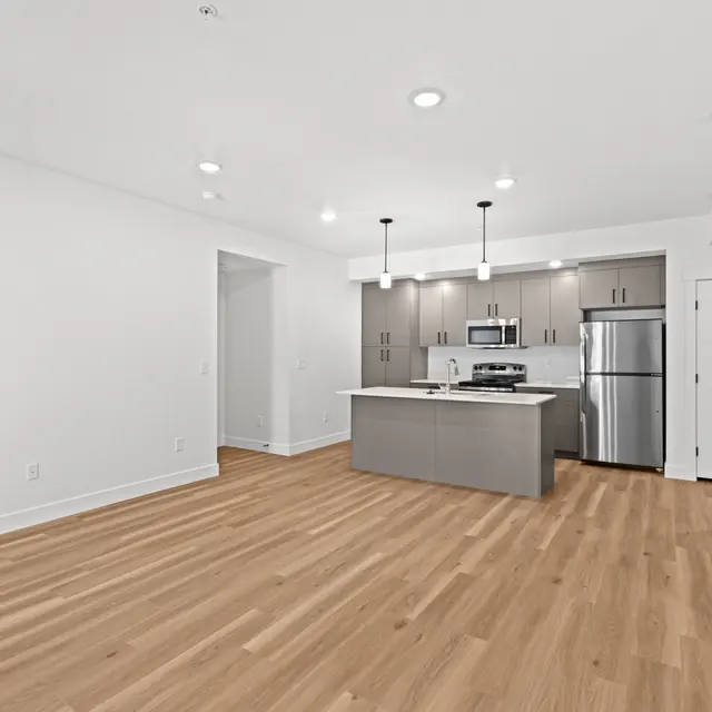 A spacious and modern apartment interior featuring a kitchen with stainless steel appliances and a large island, hardwood flooring, and white walls.