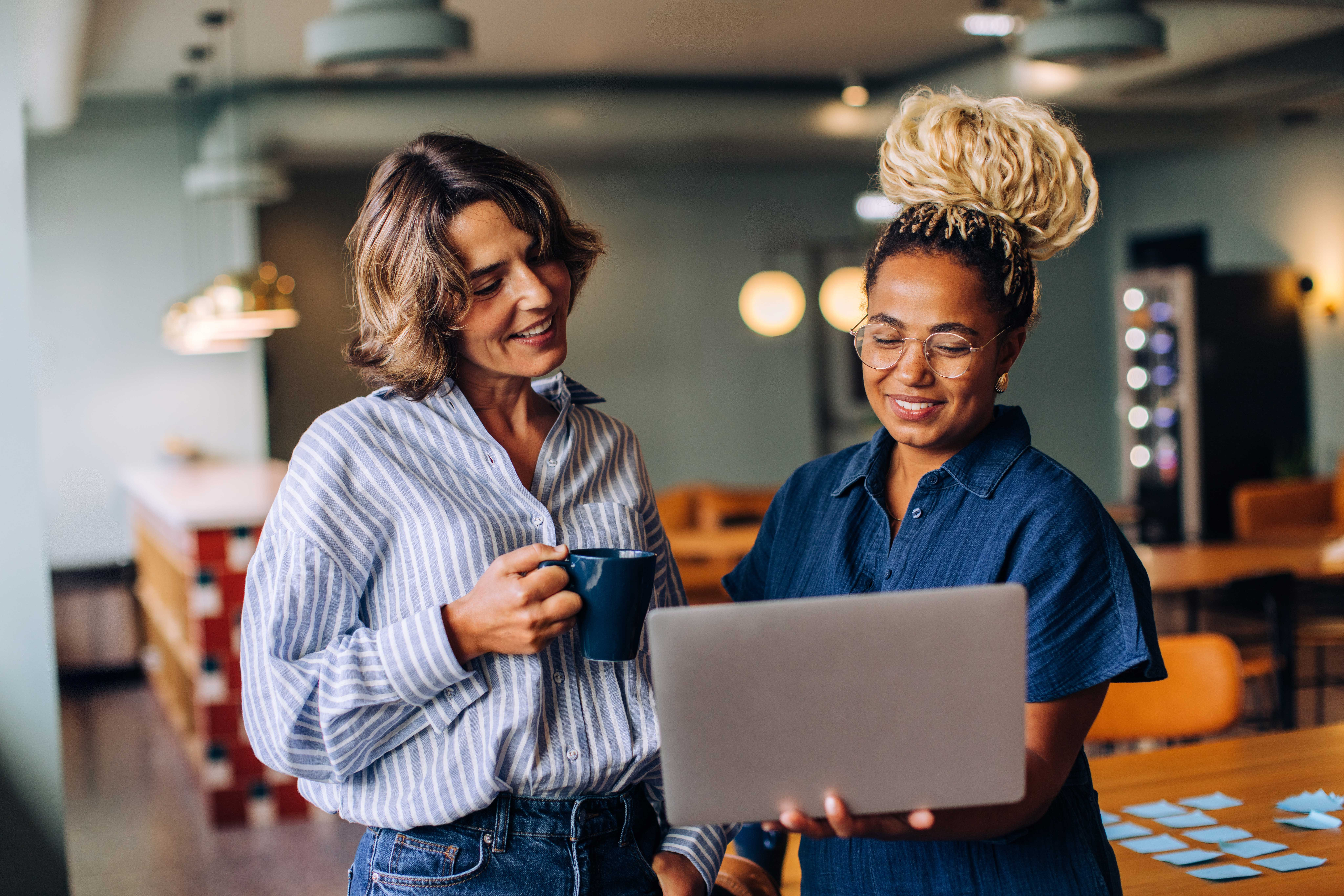 Two women discussing something on a laptop while one holds a cup of coffee in a modern workspace.