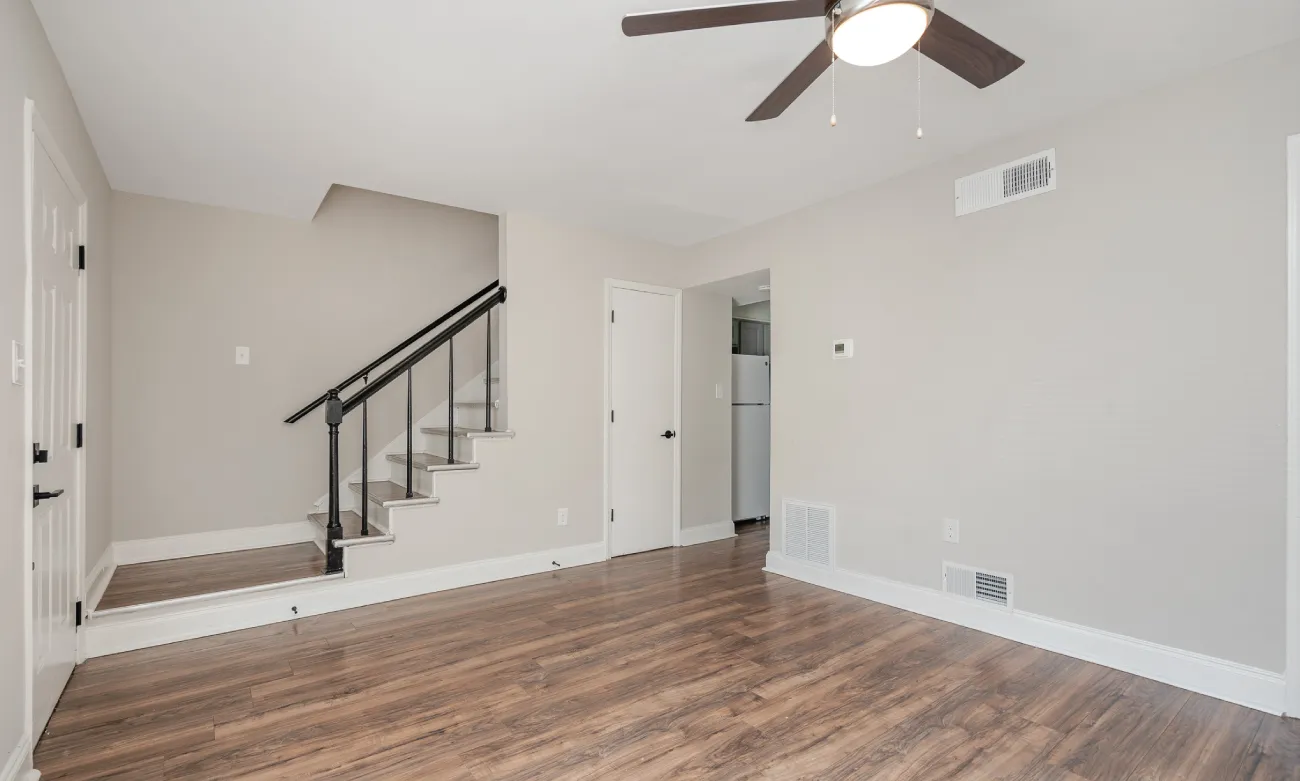 Spacious Living Room A spacious living room featuring hardwood floors and a ceiling fan. A staircase with wrought iron railings is visible on the left. The room is painted in light colors, and there is an open doorway leading to another area.