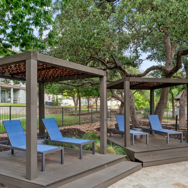 Two outdoor lounging structures with blue loungers under wooden pergolas surrounded by greenery.