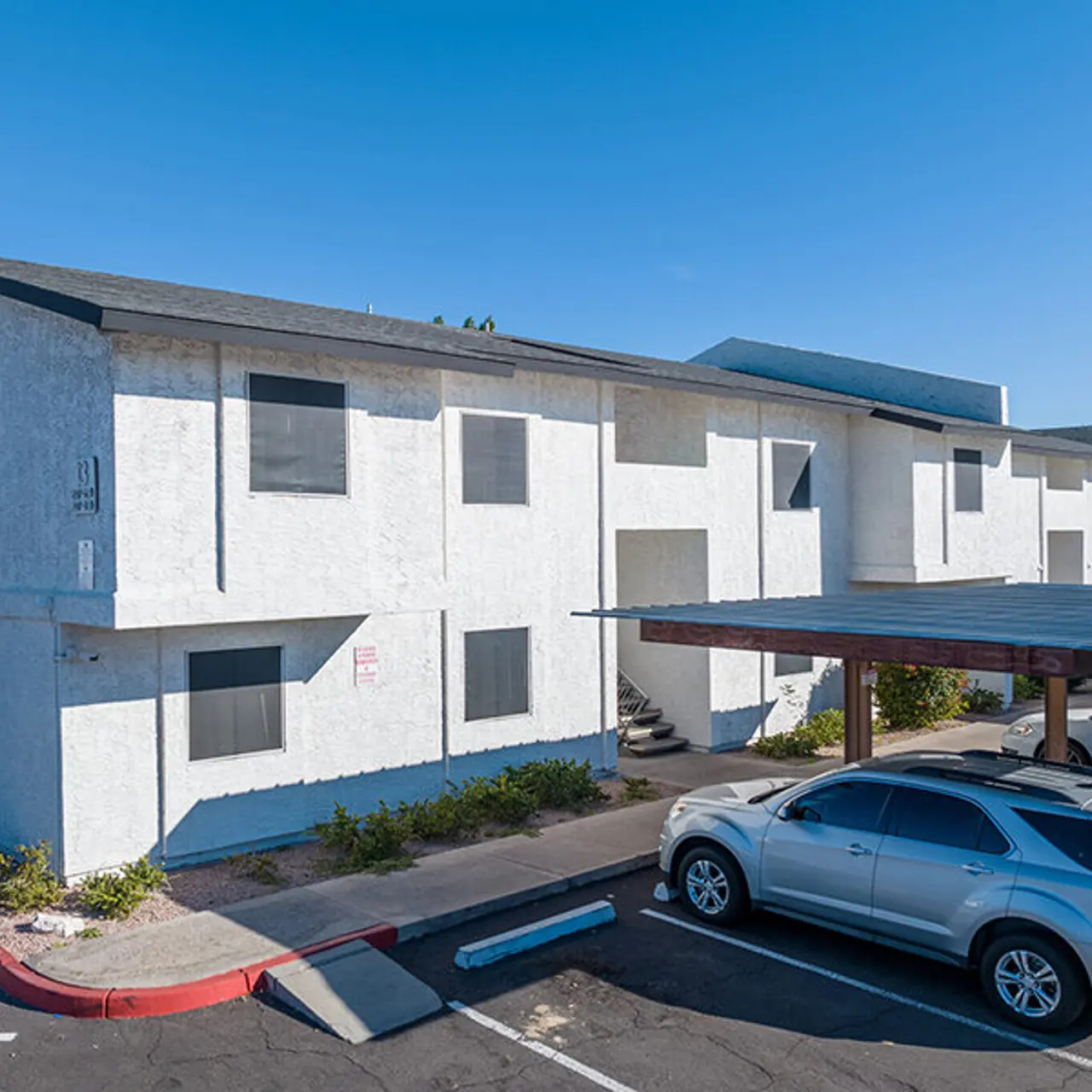 Exterior view of a two-story apartment building with a parked car in the foreground and a blue sky above.