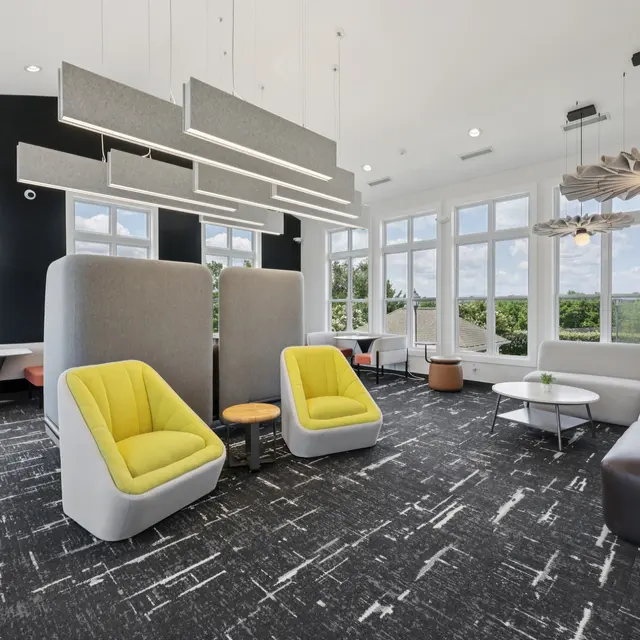 A contemporary lounge area featuring large windows, stylish furniture including a mix of gray, white, and yellow chairs, hanging light fixtures, and a carpet with a textured pattern.