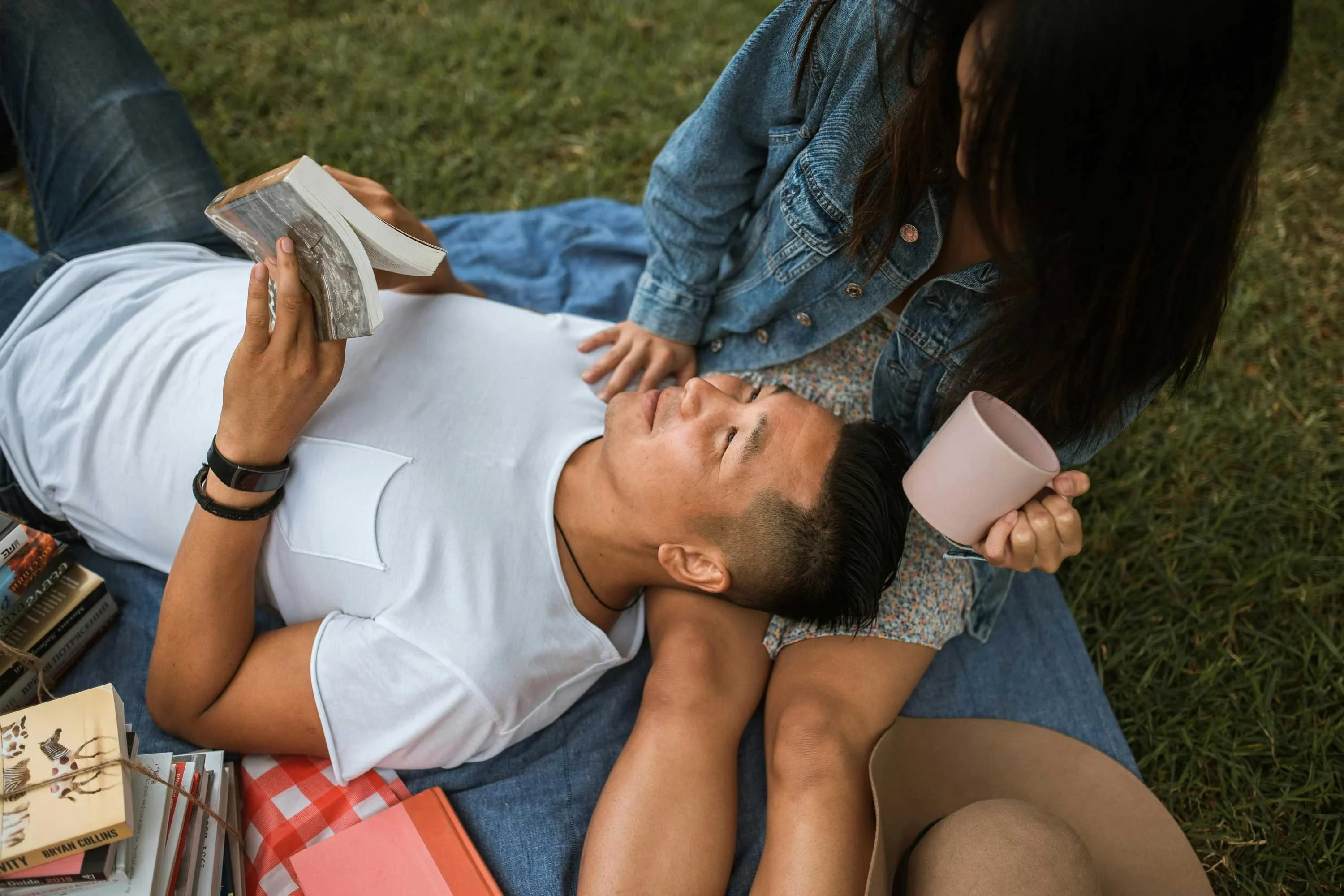 A couple enjoying a picnic on a grassy surface. One person is lying down reading a book, while the other is sitting beside them holding a coffee mug. Surrounding them are some books and a picnic blanket.