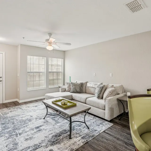 A cozy living room featuring a gray sofa with decorative cushions, a coffee table with a tray, two bright green modern chairs, a stylish rug, and large windows allowing natural light.