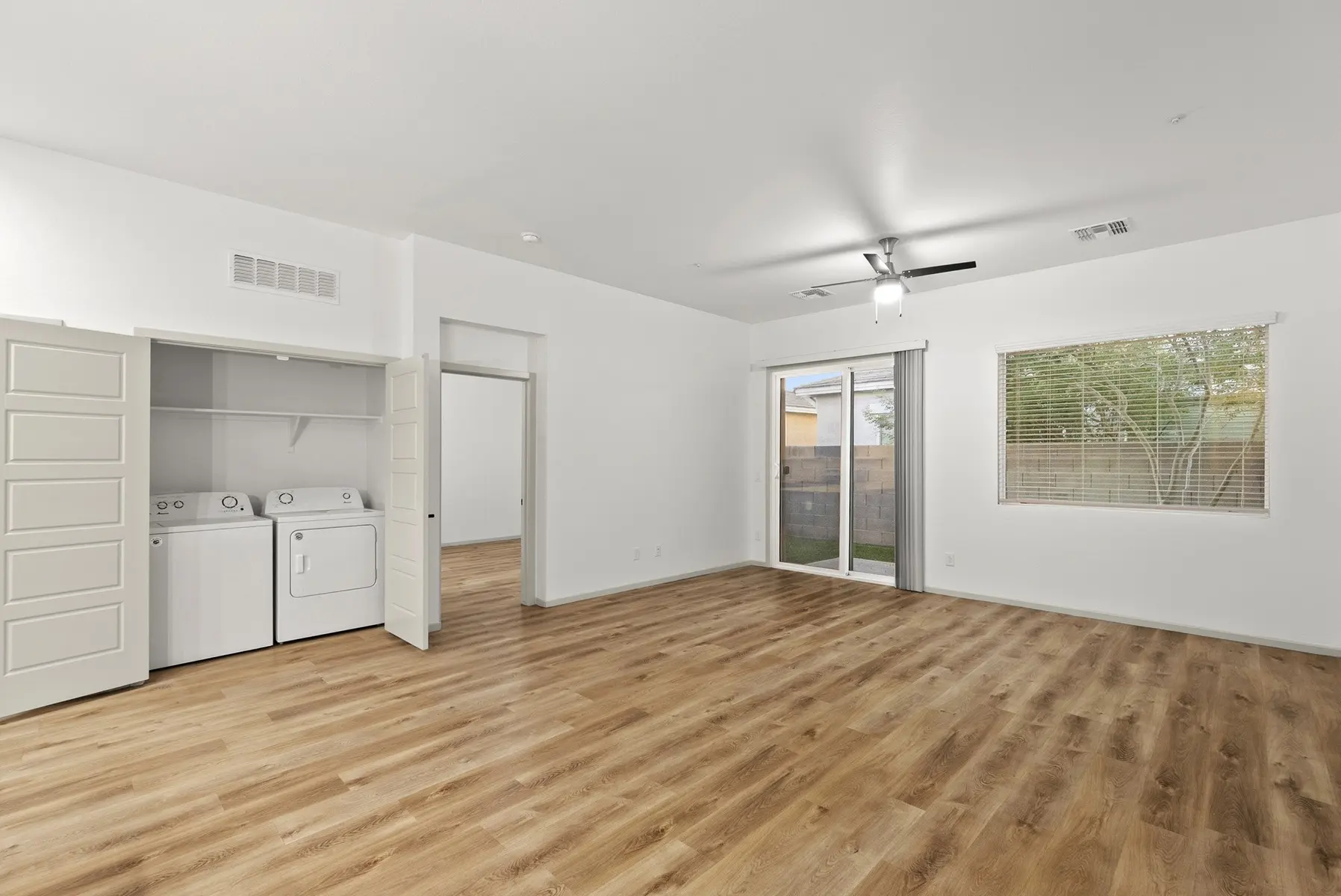 Spacious room with wood flooring, ceiling fan, laundry area, large window, and sliding glass door to outdoor space.