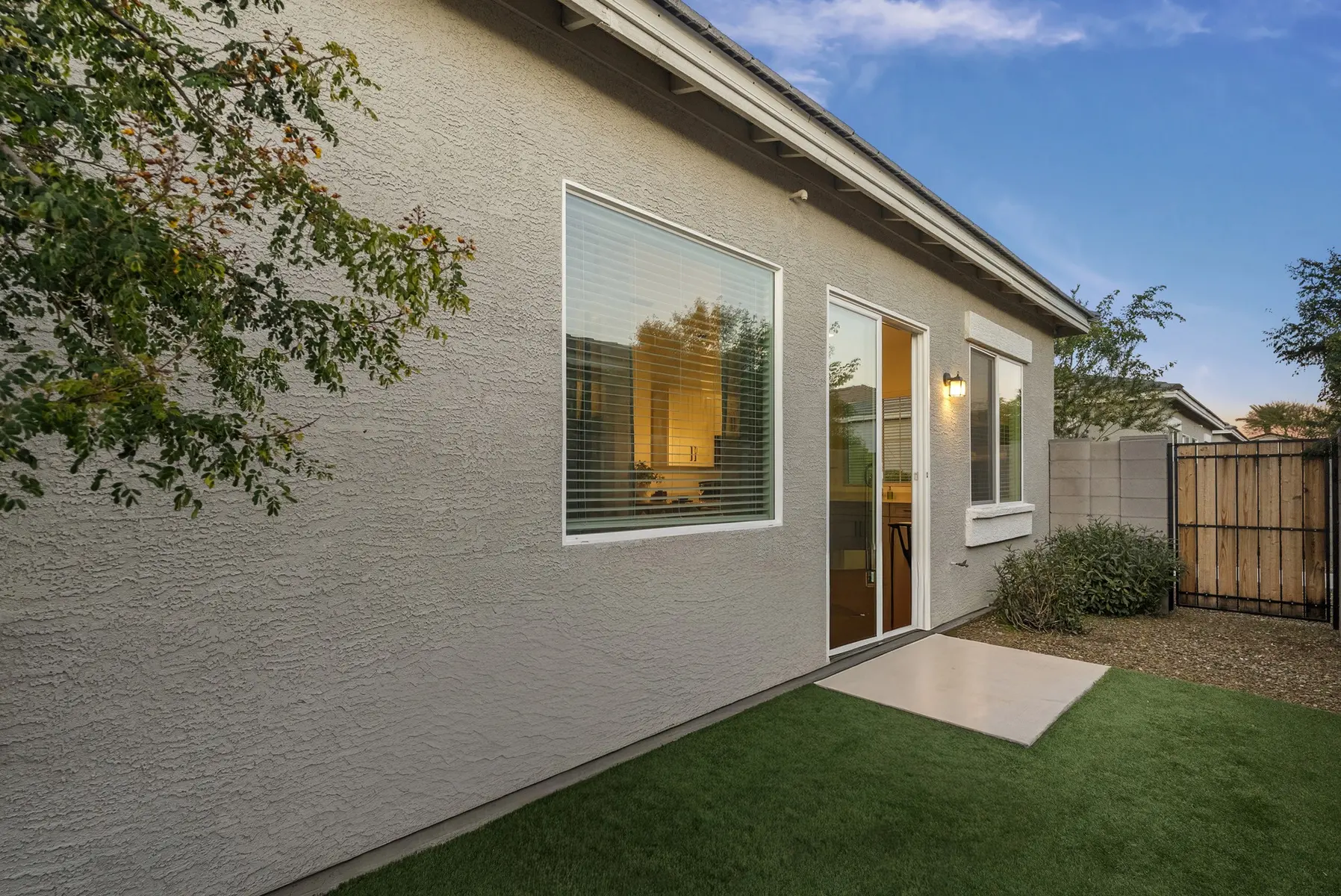Exterior view of a modern house with a patio, large window, and lawn, surrounded by shrubs and a wooden fence.