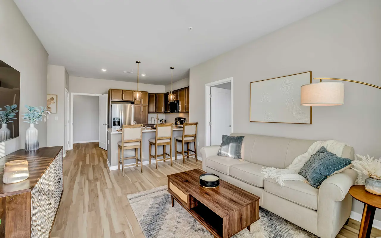 A stylish living room featuring a beige sofa, a wooden coffee table, and a partially visible kitchen area with bar stools. Light wood flooring and neutral-colored walls give it a modern feel.