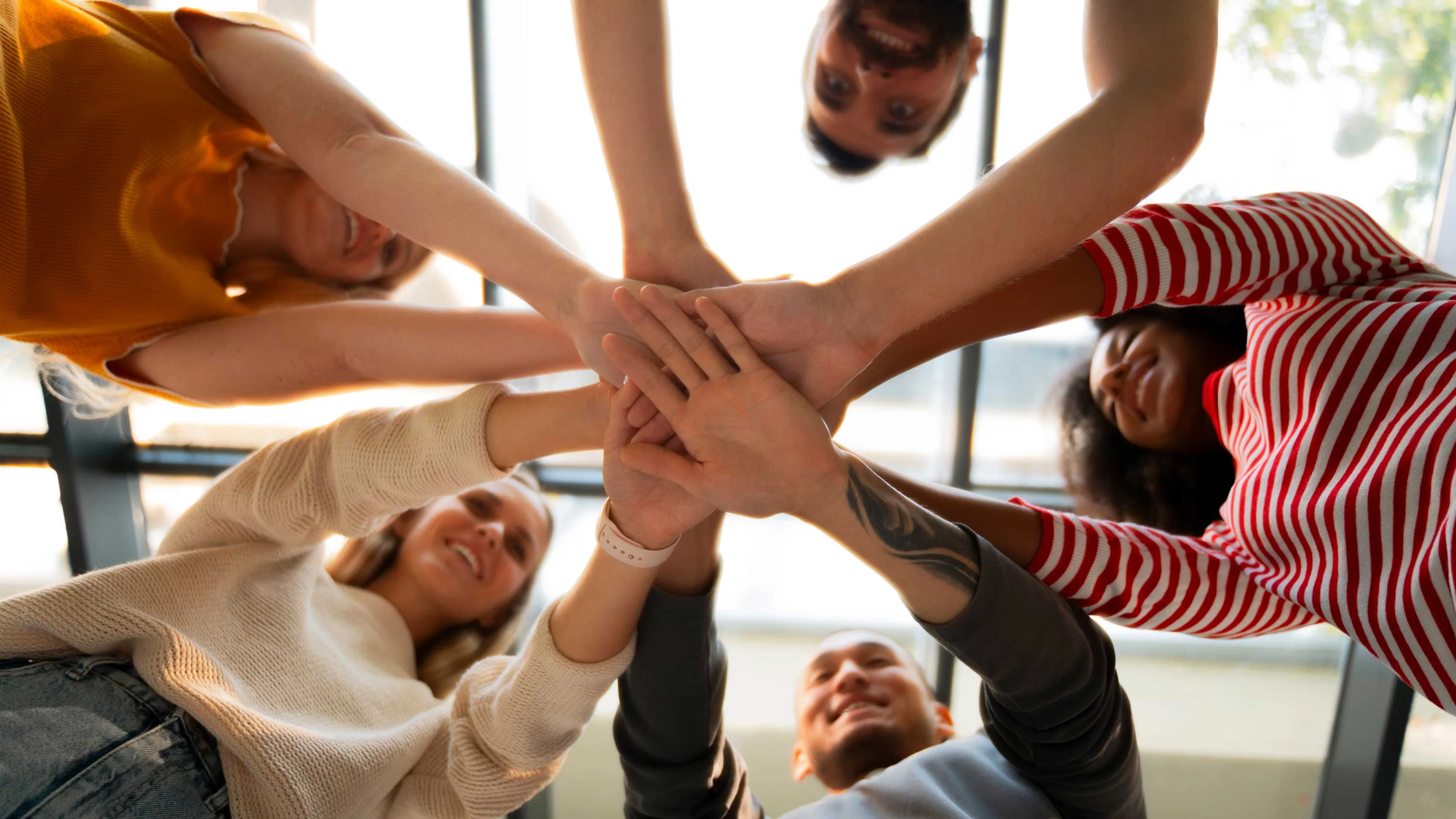 A group of five people from various angles, standing in a circle with their hands stacked together in the center. The photo is taken from an overhead perspective, showing a mix of skin tones and styles, with the background featuring large windows letting in natural light.