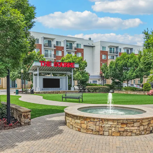 A landscaped outdoor area featuring a stone fountain in the foreground, surrounded by greenery and seating. In the background, a modern building with a sign reading 'THE GROVE'.
