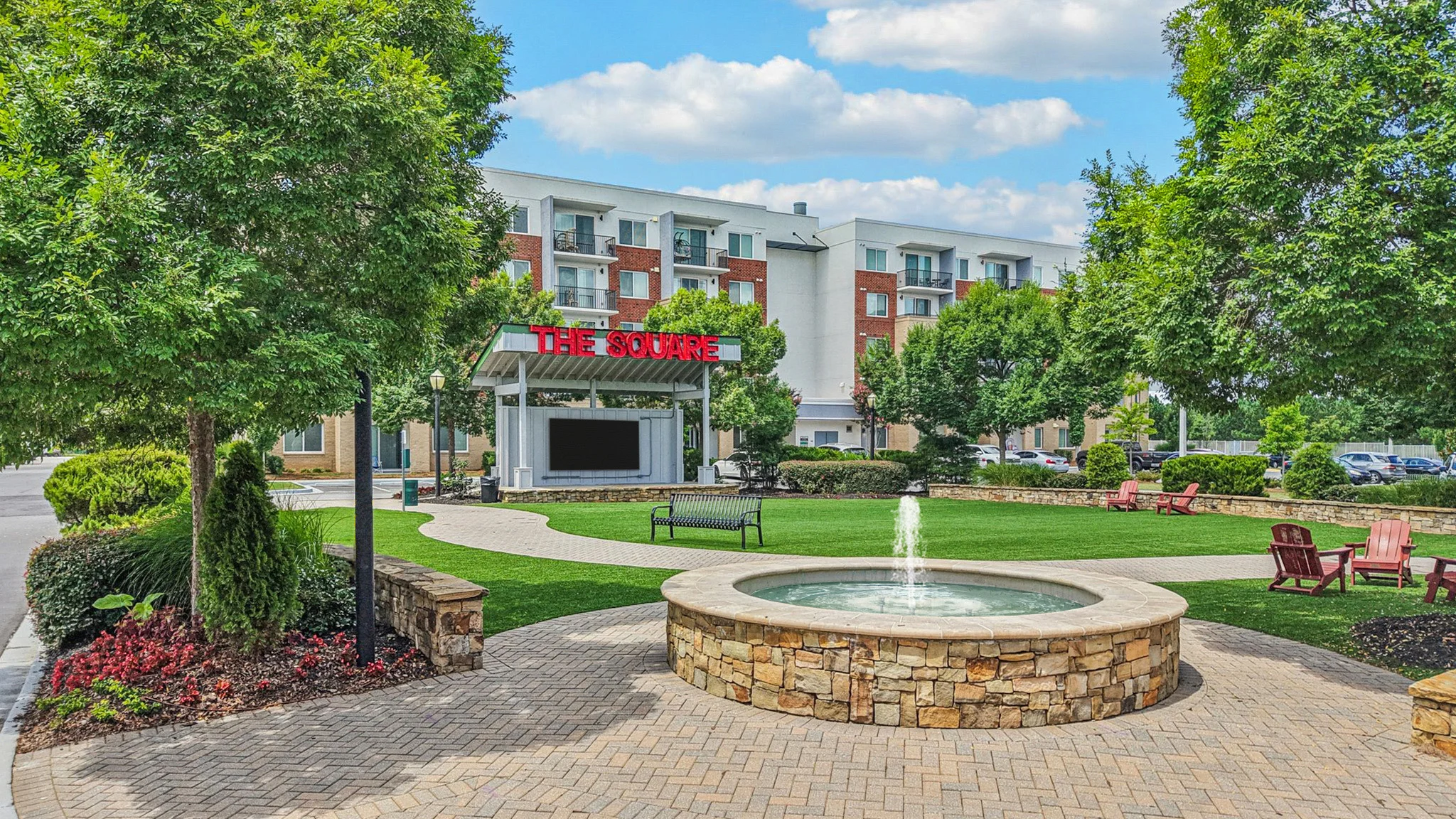 The Grove Outdoor Area A landscaped outdoor area featuring a stone fountain in the foreground, surrounded by greenery and seating. In the background, a modern building with a sign reading 'THE GROVE'.