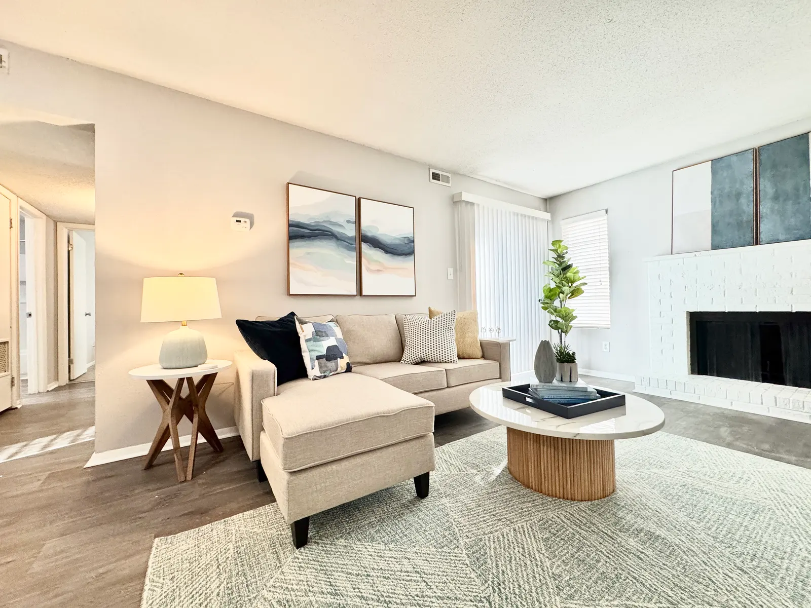 A cozy living room featuring a light-colored sofa with decorative pillows, a round coffee table, and a floor lamp. There are abstract paintings on the wall, large windows with blinds, and a modern white fireplace.