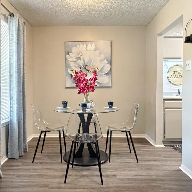 A modern dining area featuring a round glass table with four transparent chairs, a floral painting on the wall, and a vase of pink orchids as a centerpiece. The room is well-lit with sheer curtains and has a cozy ambiance.