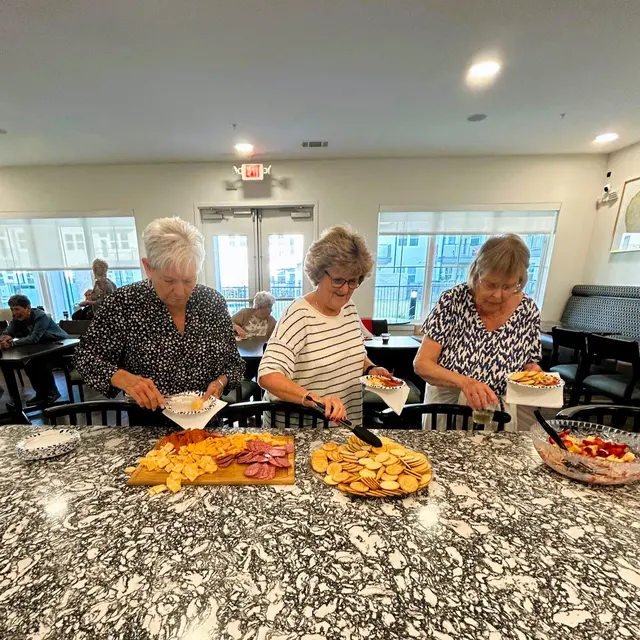 Three women preparing snacks at a community Wine Down Wednesday gathering, with a variety of chips and fruits arranged on a table.
