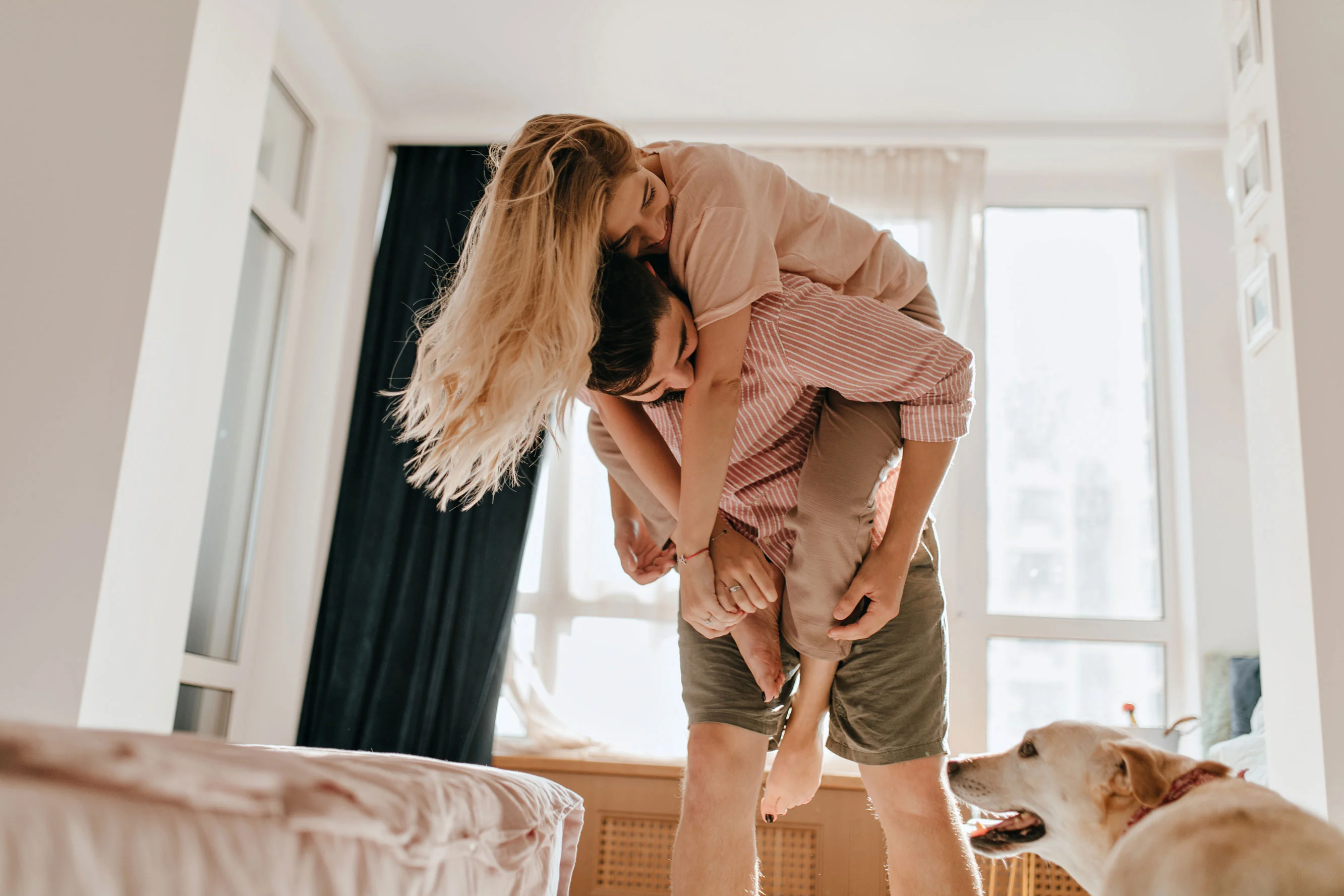 A couple playfully interacting in a bright room, with the woman giving the man a piggyback ride. A dog is partially visible in the foreground, adding to the domestic scene.