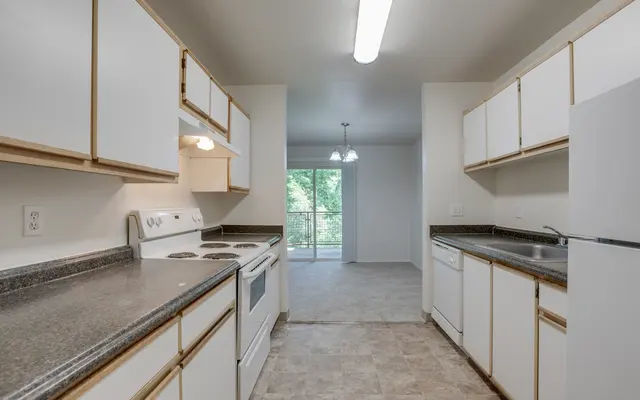 A kitchen with white cabinets and dark countertops, featuring a stove, dishwasher, and a view of a light-filled room with sliding doors.
