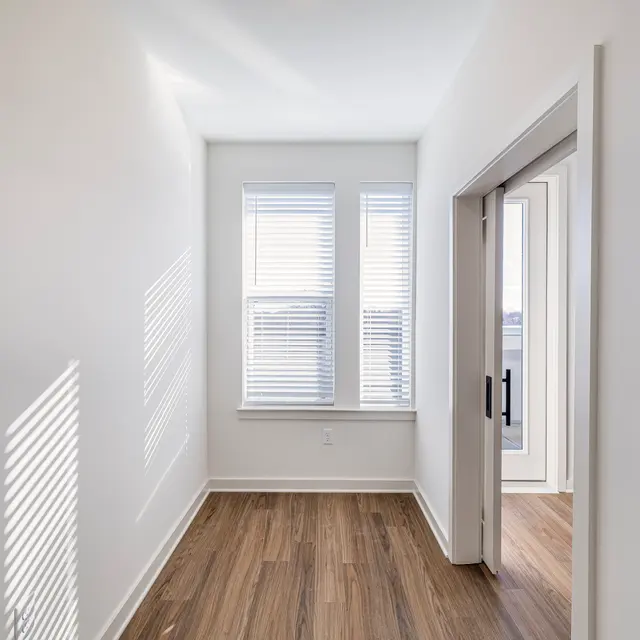 A well-lit hallway in a modern apartment featuring light-colored walls and wooden flooring. There are two windows with blinds allowing natural light to stream in, creating shadows on the wall.