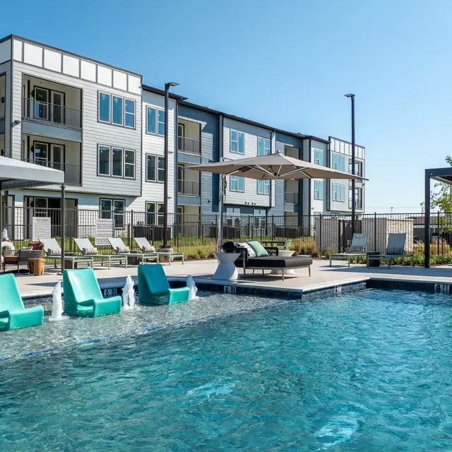 A modern apartment complex with a swimming pool in the foreground, featuring green lounge chairs by the water, shady umbrellas, and stylish buildings in the background.