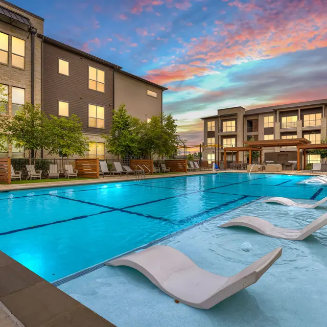 A luxurious pool area at sunset featuring lounge chairs by the poolside and modern apartment buildings in the background.