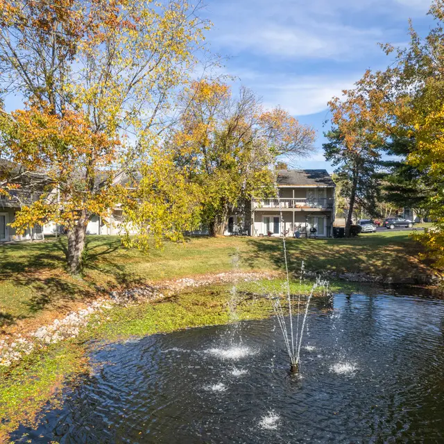 A peaceful pond with a fountain, surrounded by trees with colorful autumn leaves and a building in the background.