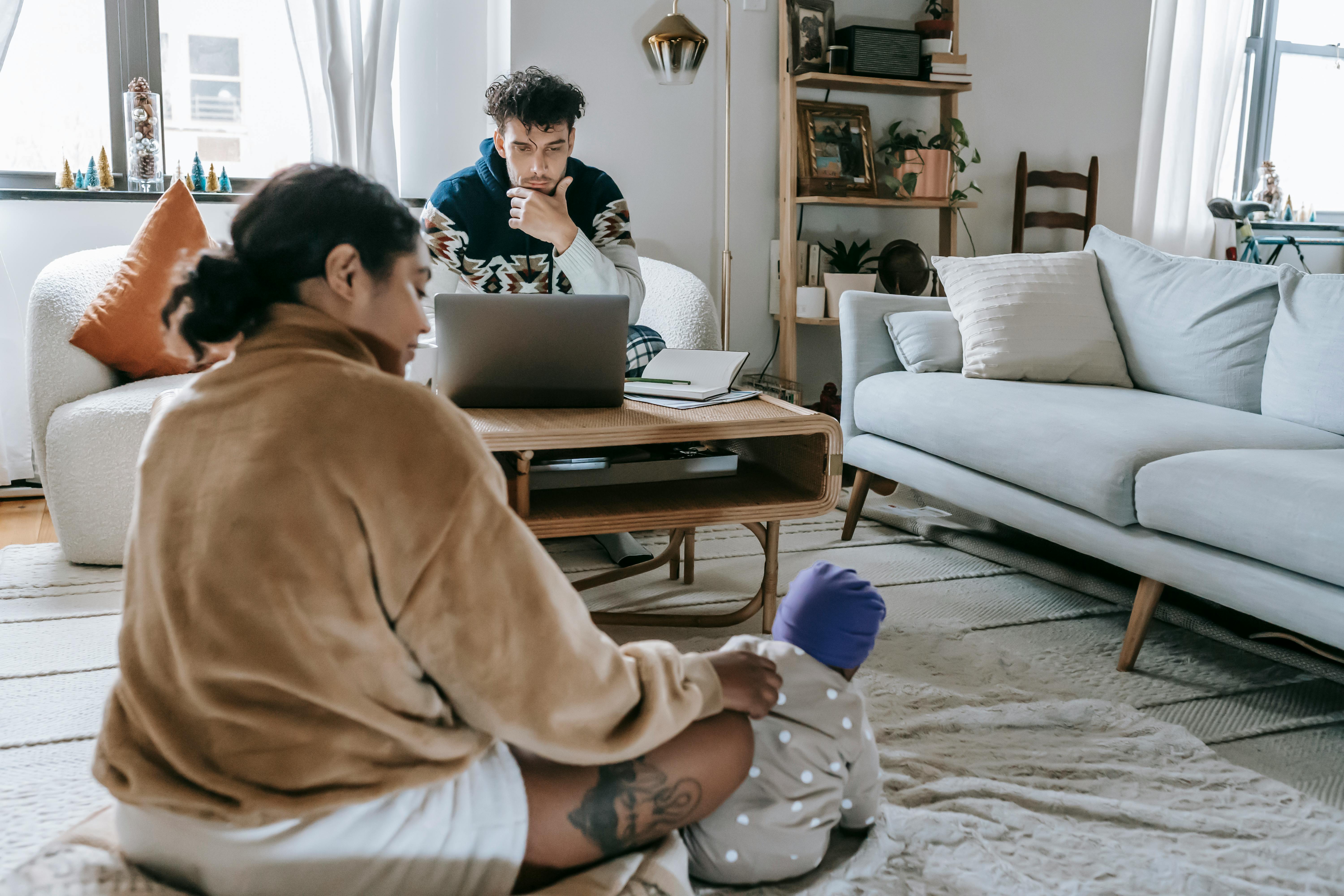 A cozy living room scene with a woman sitting on the floor wearing pajamas and a robe, looking at her legs, while a man sits on a couch in the background with a laptop.