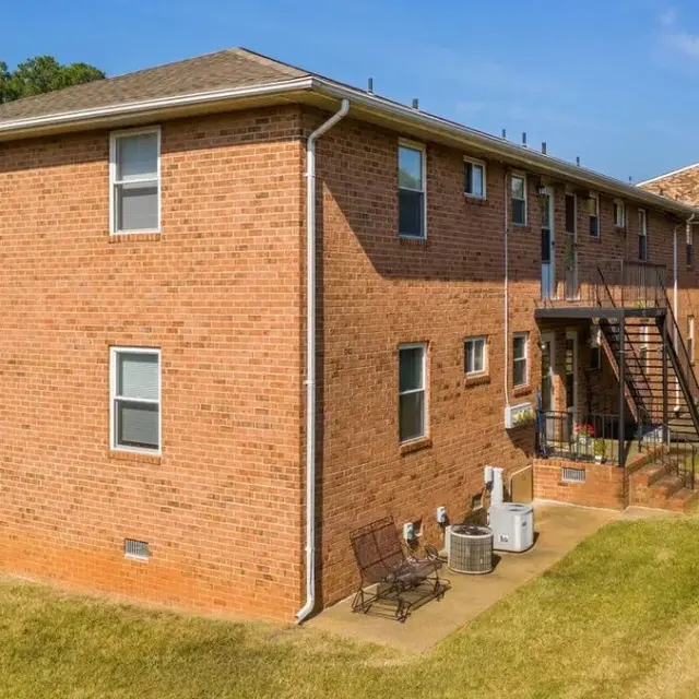 Exterior view of a red brick apartment building featuring a lawn, stairs, and multiple windows.