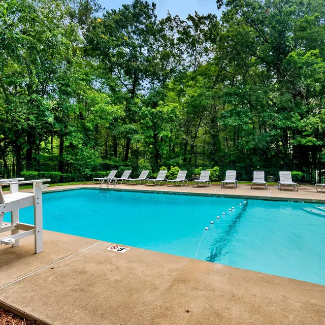 A serene swimming pool area surrounded by lush greenery. The pool is clear and inviting, with lounge chairs arranged neatly around it. A lifeguard chair is positioned at the edge of the pool.
