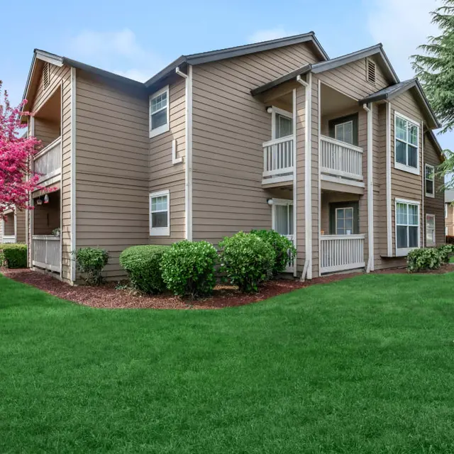 A two-story apartment building in a landscaped courtyard with green lawn and pink flowering trees.