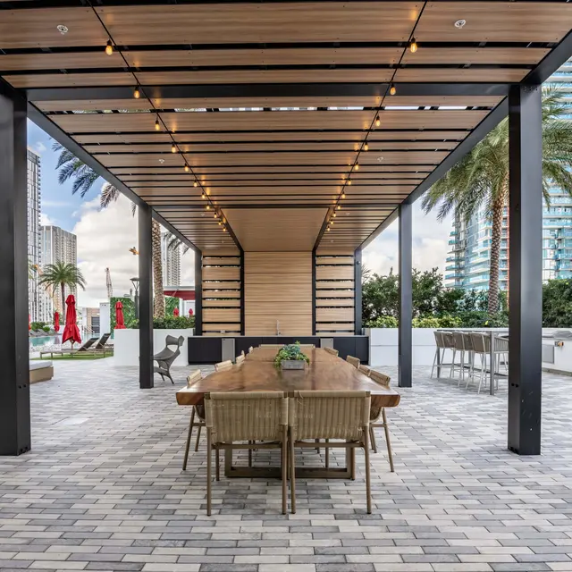 An outdoor dining area with a long wooden table and chairs under a wooden pergola. The floor is paved with blocks, and palm trees and tall buildings can be seen in the background.