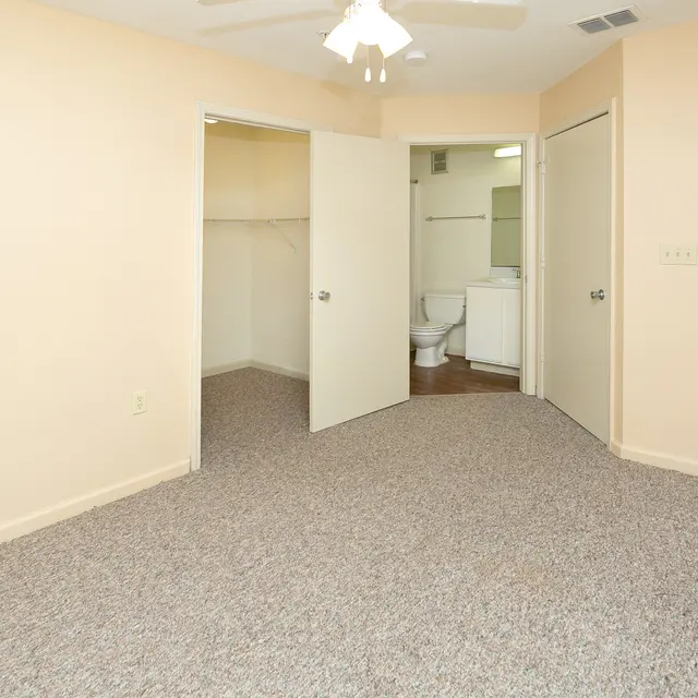 A spacious room with beige walls and carpet, featuring two doors leading to other areas and a ceiling fan.