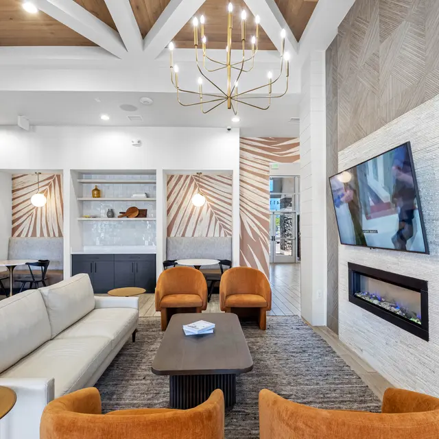 A modern living room featuring a comfortable sectional sofa, accent chairs, and a coffee table in front of a wall-mounted television and a fireplace. The ceiling has decorative beams, and the walls are adorned with a chic design. Ambient lighting creates a cozy atmosphere.