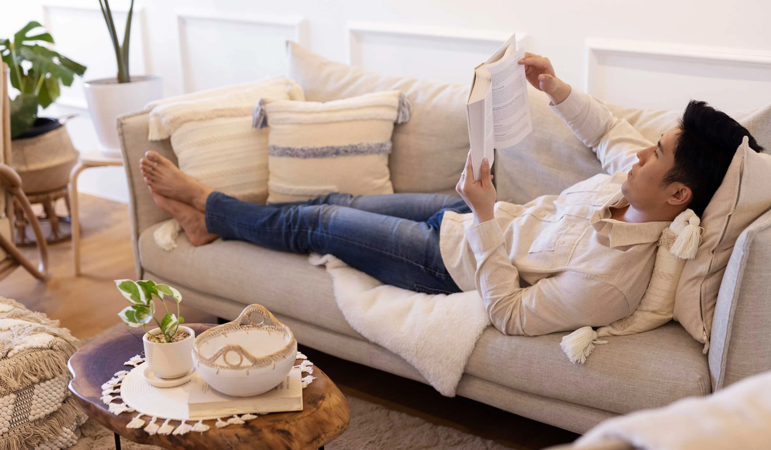 Relaxing with a Book A person lounging on a sofa while reading a book, surrounded by cozy decor, plants, and a small table with a decorative bowl and plant.