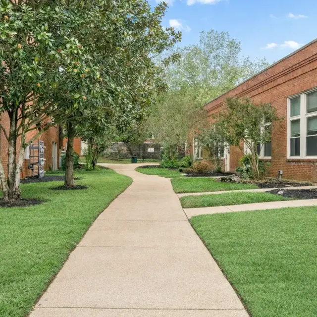 A smooth walkway lined with grass and trees, leading to a series of brick buildings under a clear blue sky.