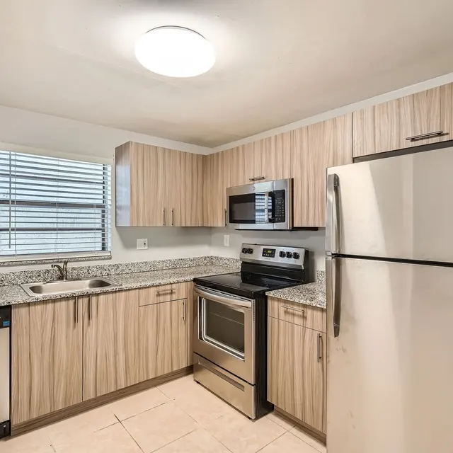 A modern kitchen featuring wooden cabinetry, granite countertops, stainless steel appliances including an oven, microwave, dishwasher, and refrigerator, with a window allowing natural light.