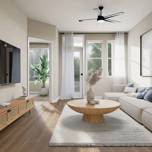 A contemporary living room featuring a light-colored sofa, a round wooden coffee table, and a television mounted on a wooden media console. Large windows with sheer curtains allow natural light to fill the space, and a potted plant adds a touch of greenery.