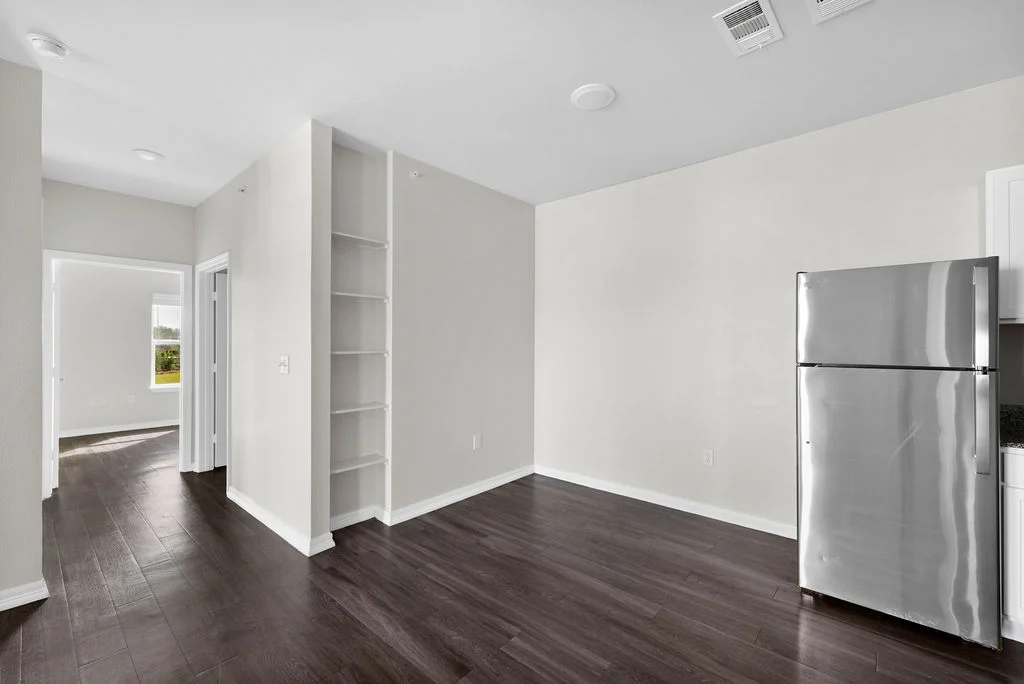 A modern kitchen and living area featuring dark wood flooring, a stainless steel refrigerator, and light-colored walls. There is an open layout with a small shelf on the left and a doorway leading to another room.