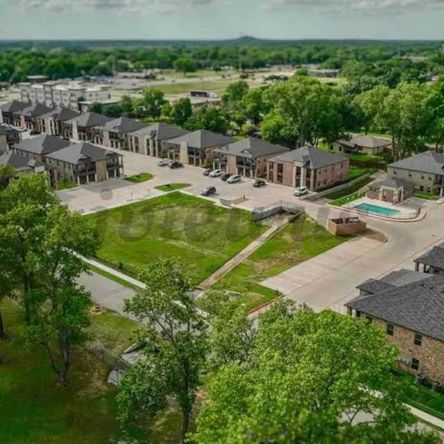 Aerial view of a residential apartment complex with multiple buildings, a swimming pool, and green trees in the surrounding area.