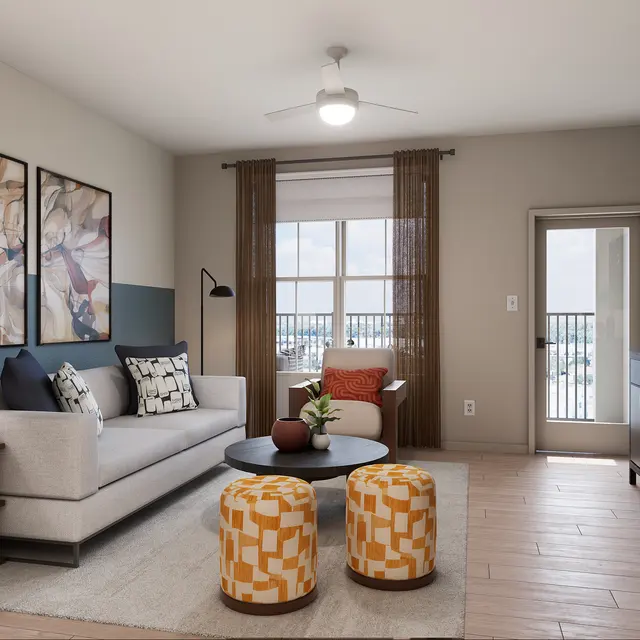 A modern living room featuring a light gray sofa with decorative pillows, orange and white patterned ottomans, a round coffee table, and a sideboard. Large windows with sheer curtains provide natural light, and artwork adorns the walls.