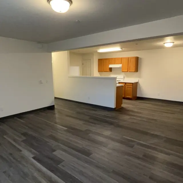 Spacious interior of a modern apartment with wood-like flooring, an open layout featuring a kitchen area with wooden cabinets and white appliances, and ample natural light.