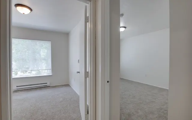 Interior view of a light-colored room with carpet, showing an open doorway leading to another room. The first room has a large window with natural light and a ceiling light fixture.