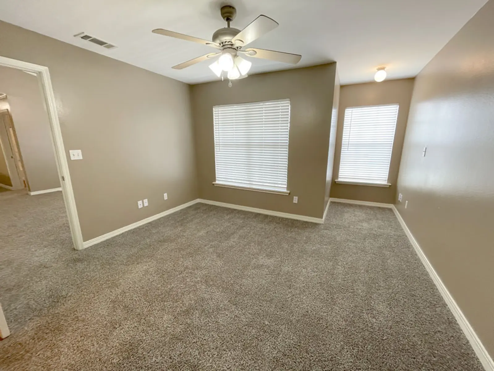 A carpeted room with light gray walls and two windows, featuring a ceiling fan.