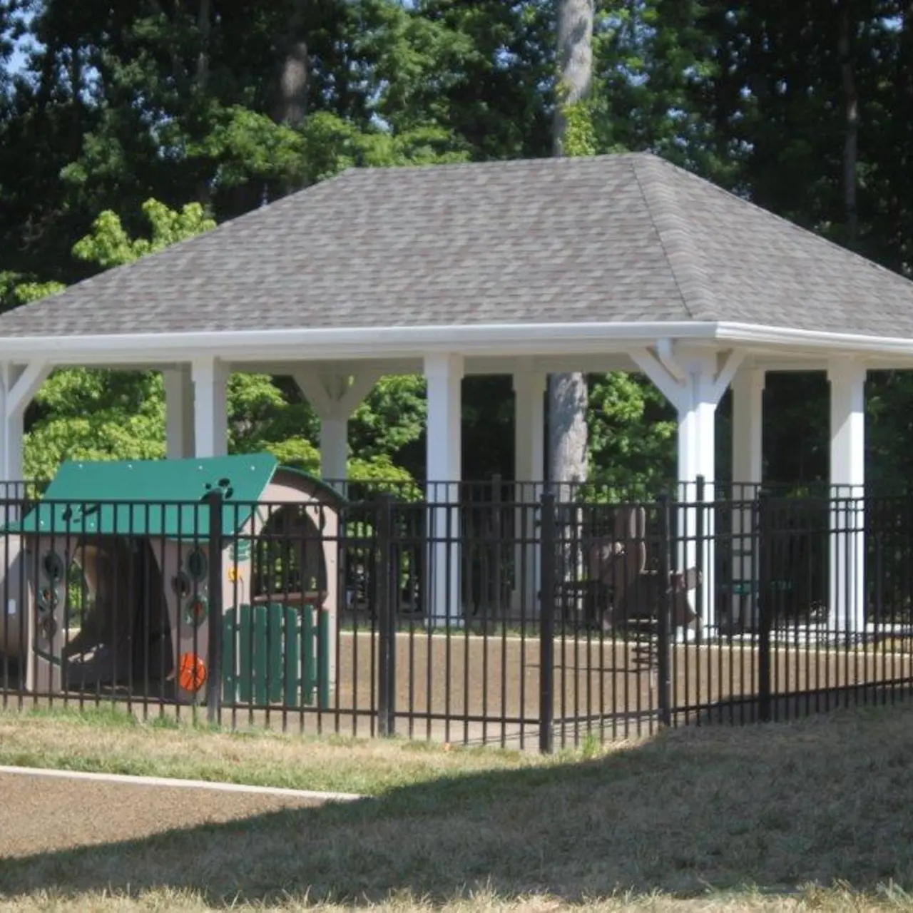 A covered pavilion structure in a playground area surrounded by a black fence and green trees in the background.