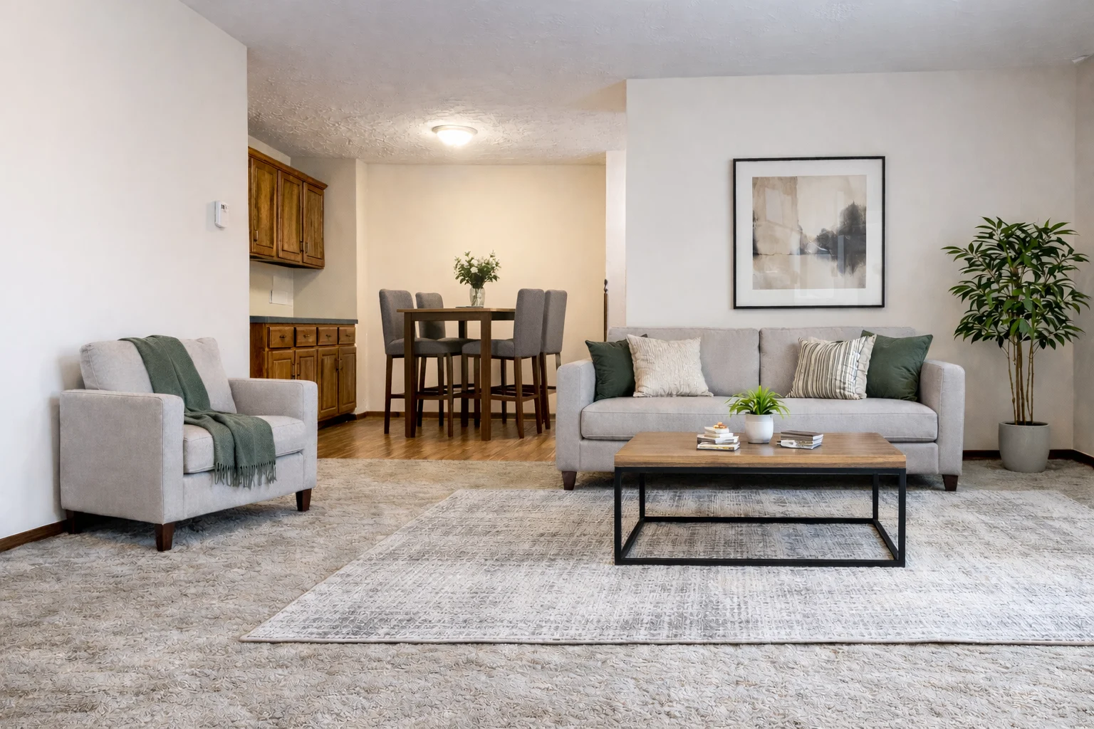 A cozy living room featuring a light gray sofa with green and white pillows, a wooden coffee table with decorative items, and a matching armchair with a green throw. In the background, there is a dining area with a small table and chairs, and a large plant in the corner. The walls are light-colored, and the flooring is carpeted with a textured area rug.