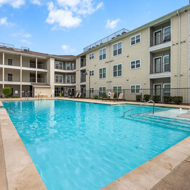 A swimming pool surrounded by apartment buildings, featuring lounge chairs and clear blue water under a partly cloudy sky.