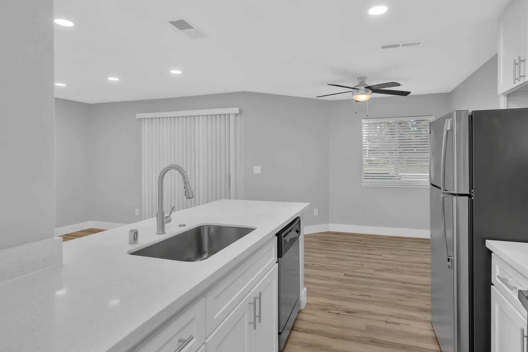 A modern kitchen interior featuring a white granite countertop, silver appliances, and a ceiling fan. The walls are painted a light gray, illuminated by recessed lighting. There is a window with blinds and wooden flooring that gives a warm look to the space.