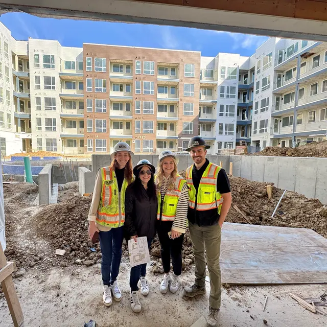 Construction Site Visit Four individuals standing in a construction site, wearing safety vests and hard hats, with a multi-story building under construction in the background.