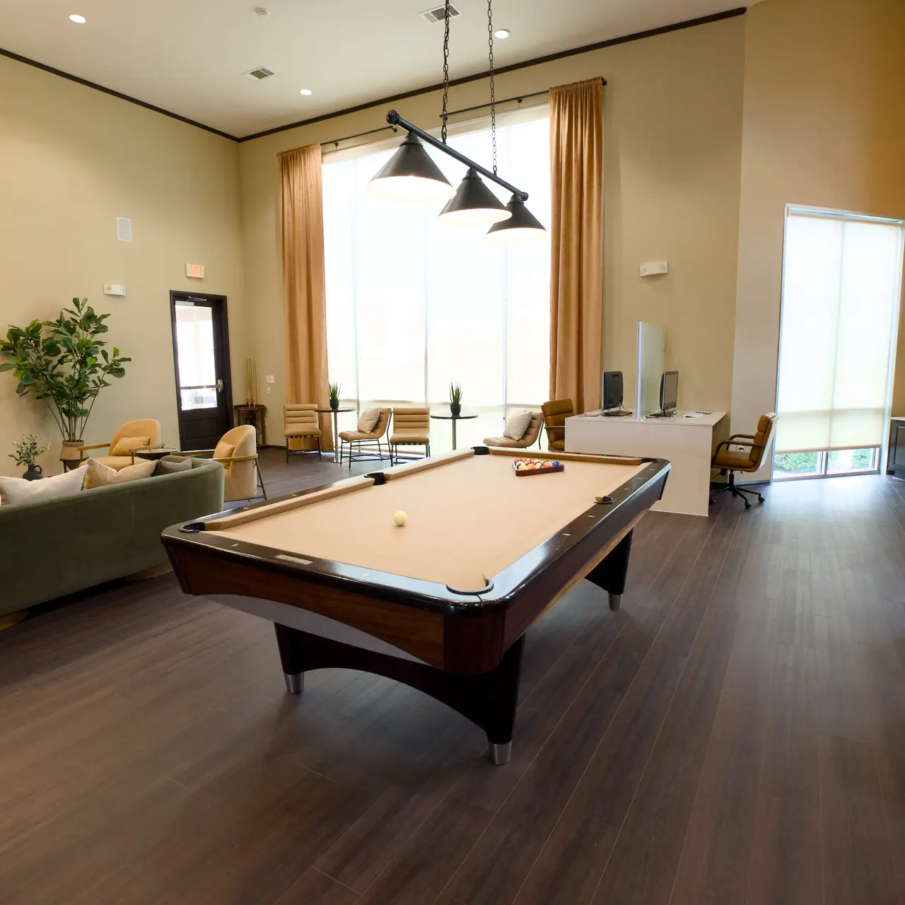 A spacious common area featuring a pool table, a sitting area with green chairs, and large windows providing natural light. A TV is mounted on the wall and there are some plants enhancing the decor.