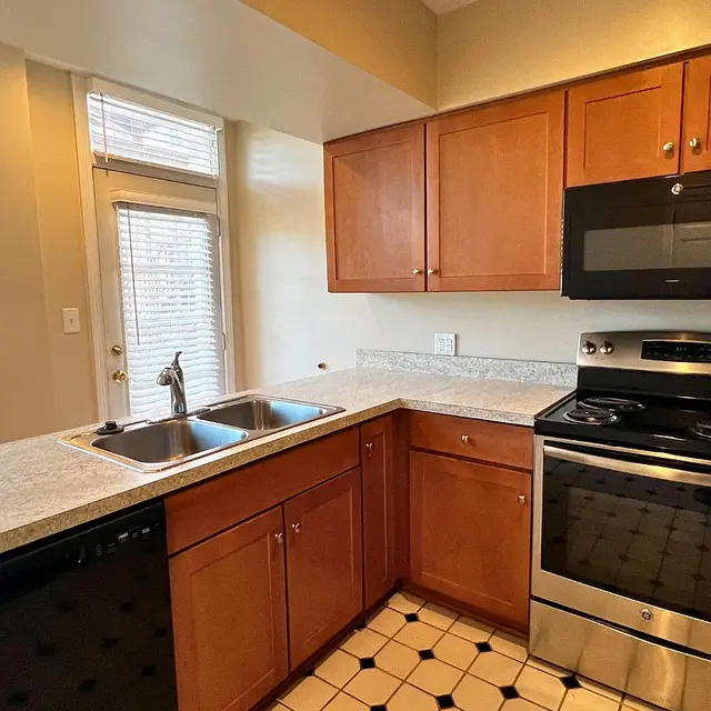 A modern kitchen featuring wooden cabinets, a stainless steel stove and microwave, a double sink, and a tiled floor, with natural light coming through the windows.