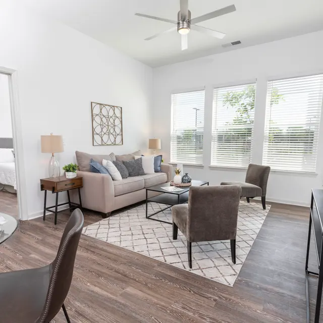 A modern living room featuring a grey couch, two chairs, and a coffee table on a patterned rug. Large windows provide natural light. A dining area with a round table is visible.