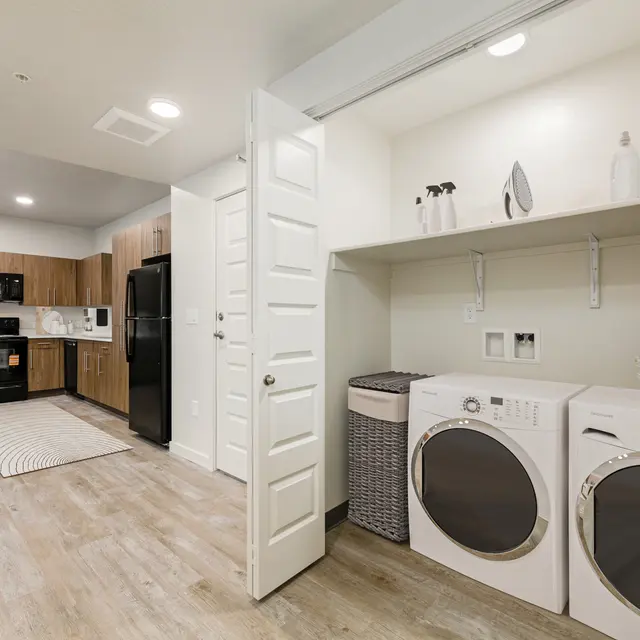 A modern laundry room featuring a washer and dryer, a large countertop, and wooden kitchen cabinets. The room has a light and airy feel with natural wood flooring.